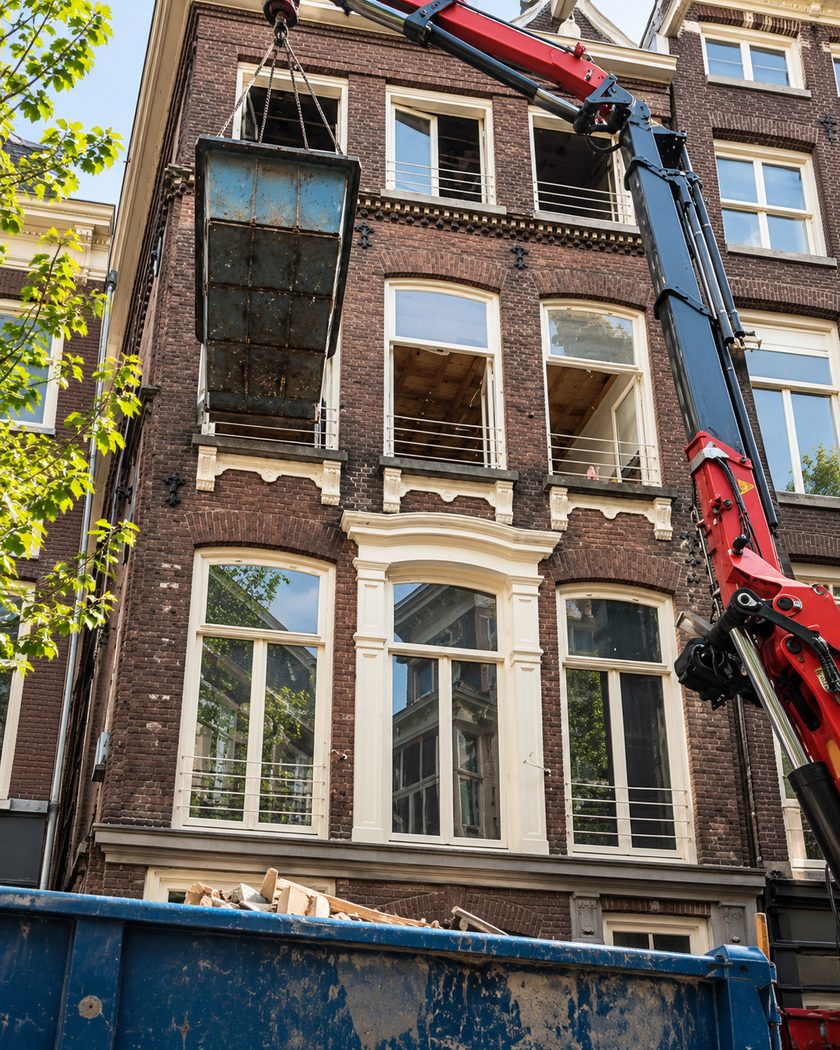 Kraan hijst een puinbak langs de gevel van een grachtenpand, met een container ervoor