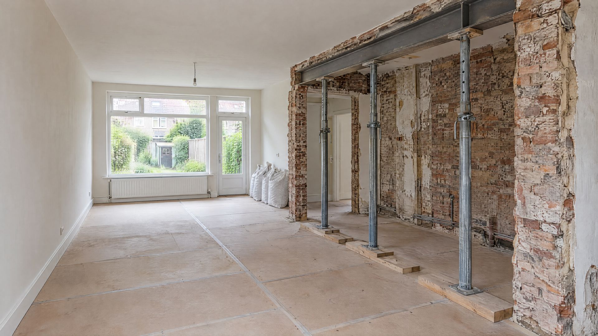 Woonkamer waar een dragende muur is weggehaald, met een stalen ligger op tijdelijke stempels en gestript metselwerk