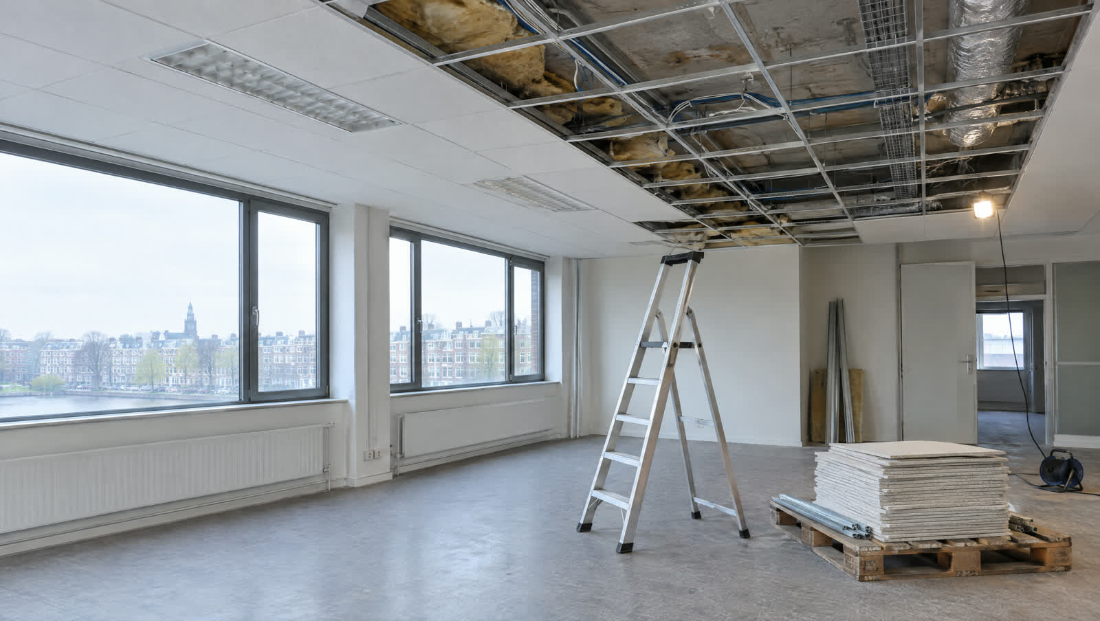 Kantoorruimte waar een systeemplafond deels is verwijderd, met het metalen raster, de betonvloer erboven en een trap